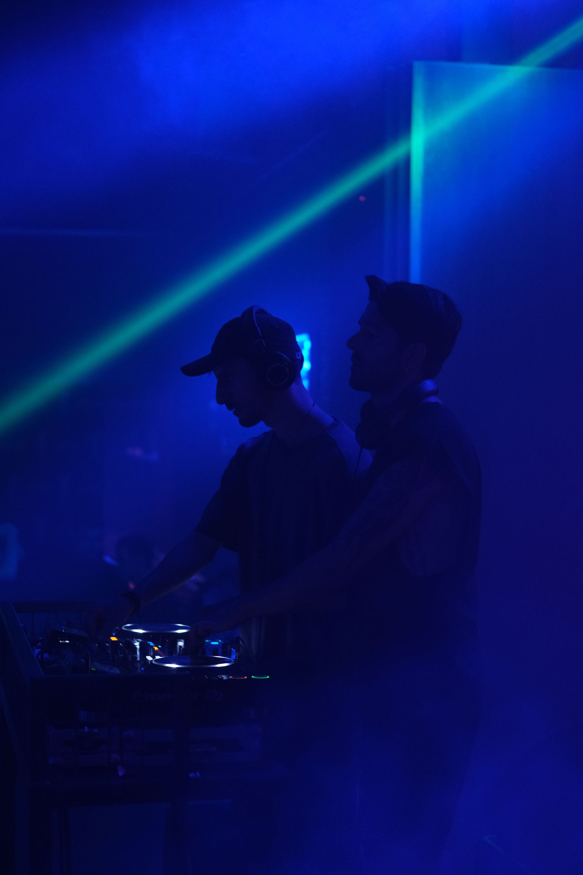 Two DJs performing live on stage in a neon-lit environment, spinning tracks with audio equipment.